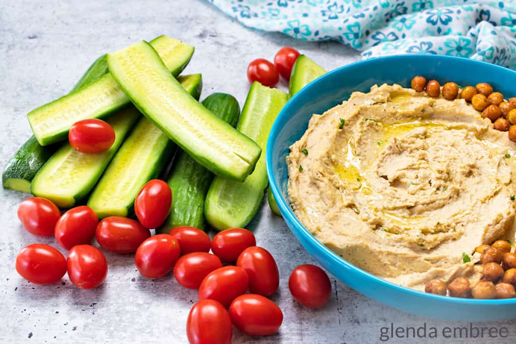 Creamy Hummus in a blue stoneware bowl next to halved cucumbers and cherry tomatoes on a concrete countertop