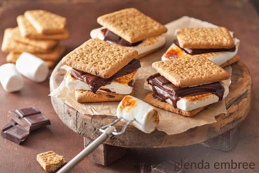 smores on a rustic wood tray with a toasted marshmallow on a metal fork