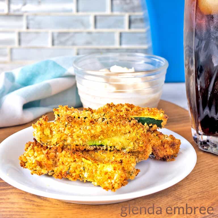 Crispy Air Fryer Zucchini on a white plate that is resting on a wooden cutting board with a bowl of Creamy Chipotle Parmesan Aioli, a glass of soda and a blue print fabric napkin