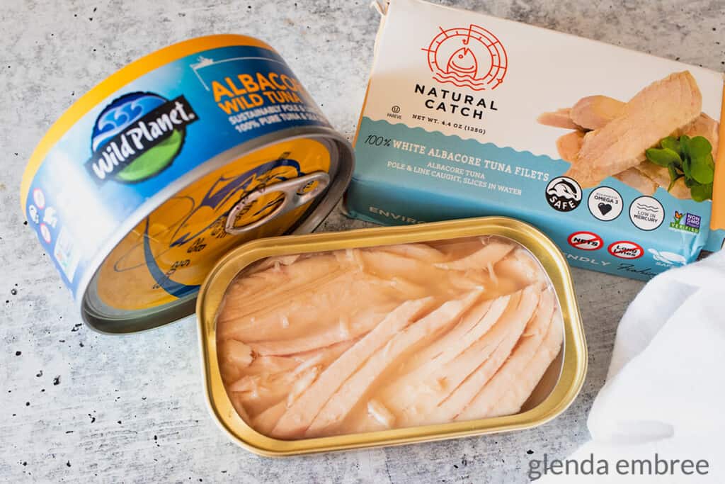 two different brands of canned tuna on a concrete counter top with an open tin of tuna in front