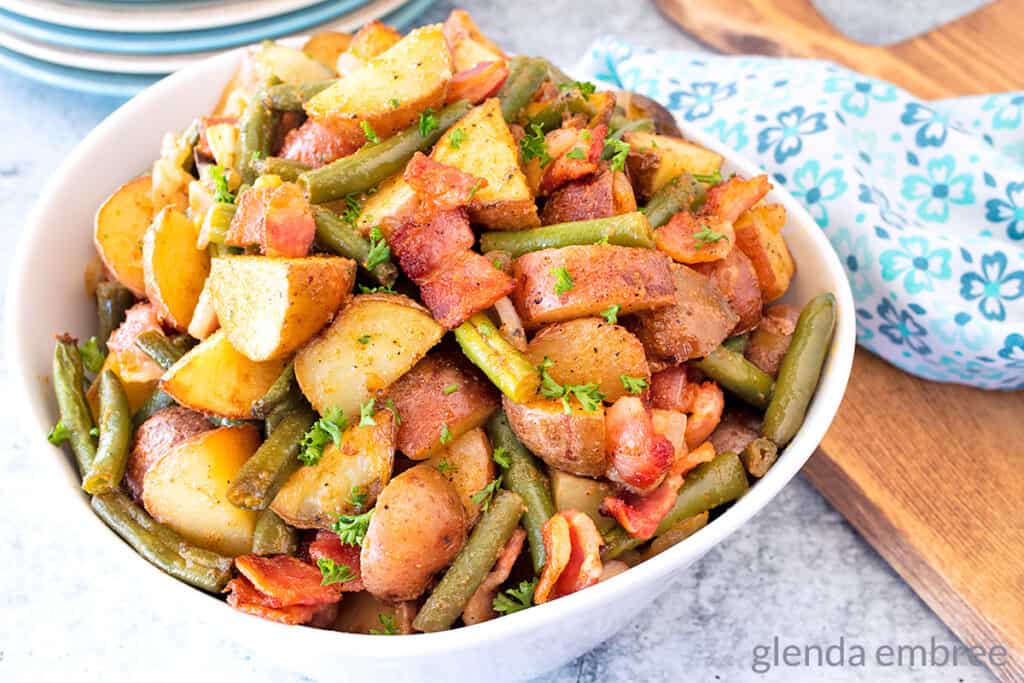 Green Beans and Potatoes in a white serving bowl on table with stack of plates and a blue print cloth napkin