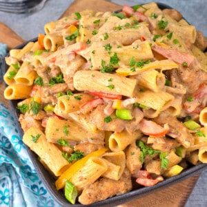 Rasta Pasta - chicken casserole - on a wooden cutting board that is sitting on a concrete counter with a stack of plate and a blue print fabric napkin