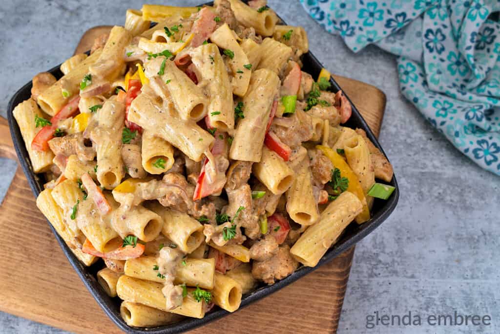 Rasta Pasta - chicken casserole - on a wooden cutting board that is sitting on a concrete counter with a stack of plate and a blue print fabric napkin