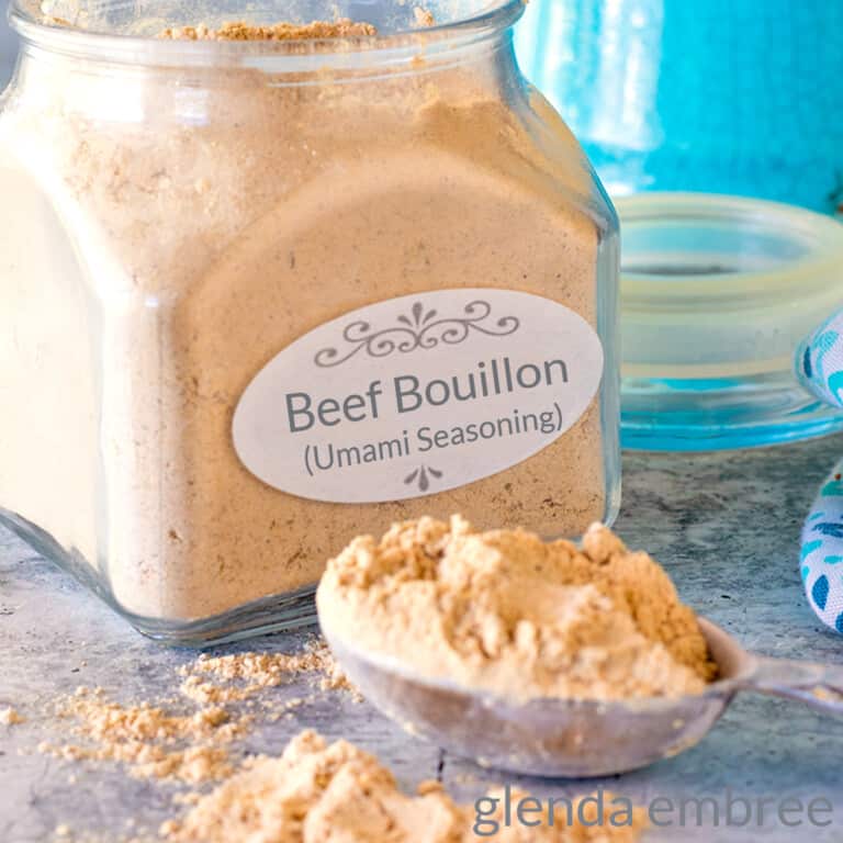 homemade beef bouillon in an apothecary jar on a concrete counter. A heaping Tablespoon of bouillon sits on the counter in front of the jar.