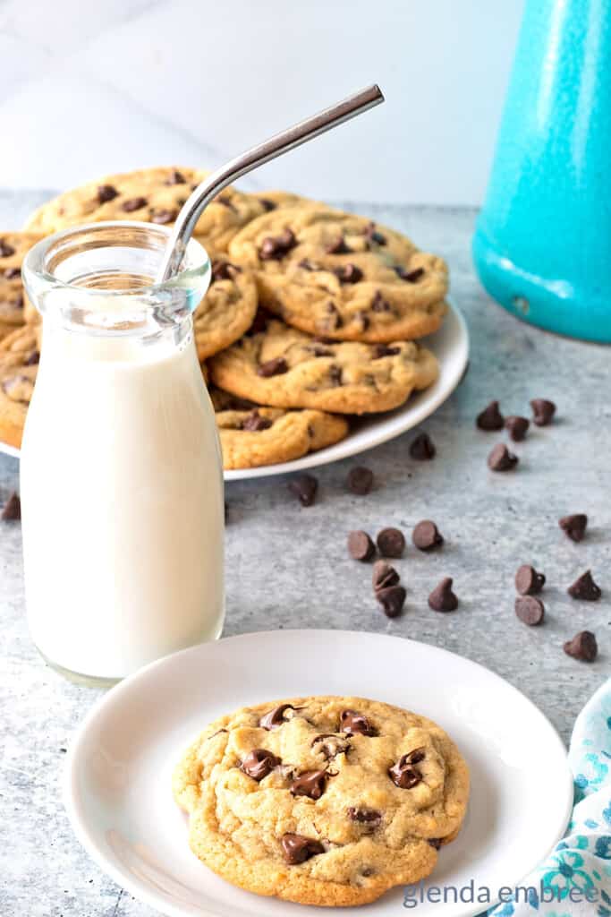 One Small Batch Chocolate Chip Cookie on a white plate with blue print napkin and bottle of milk and a plate of cookies in the background