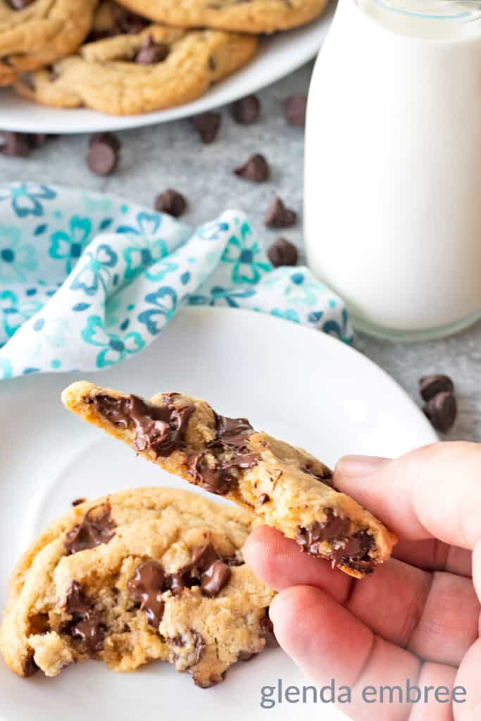 One Small Batch Chocolate Chip Cookie on a white plate with blue print napkin and bottle of milk.  cookie is broken in half to show the soft, gooey center.