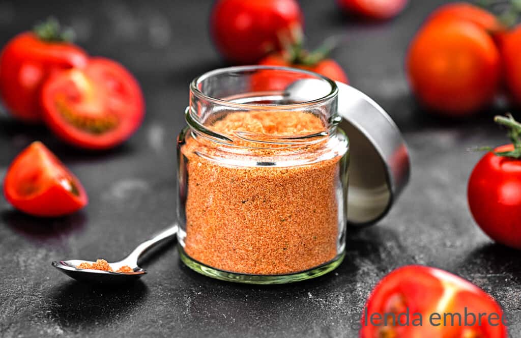 tomato powder in a glass jar, sitting on a black counter with some fresh cut tomatoes strewn about.