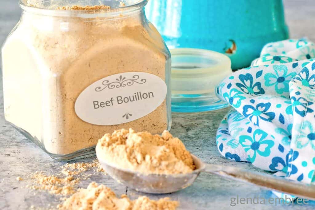 Homemade Beef Bouillon Powder in a glass apothecary jar. The lid is off and a heaping Tablespoon of Bouillon powder is sitting on the concrete countertop in front of the jar.