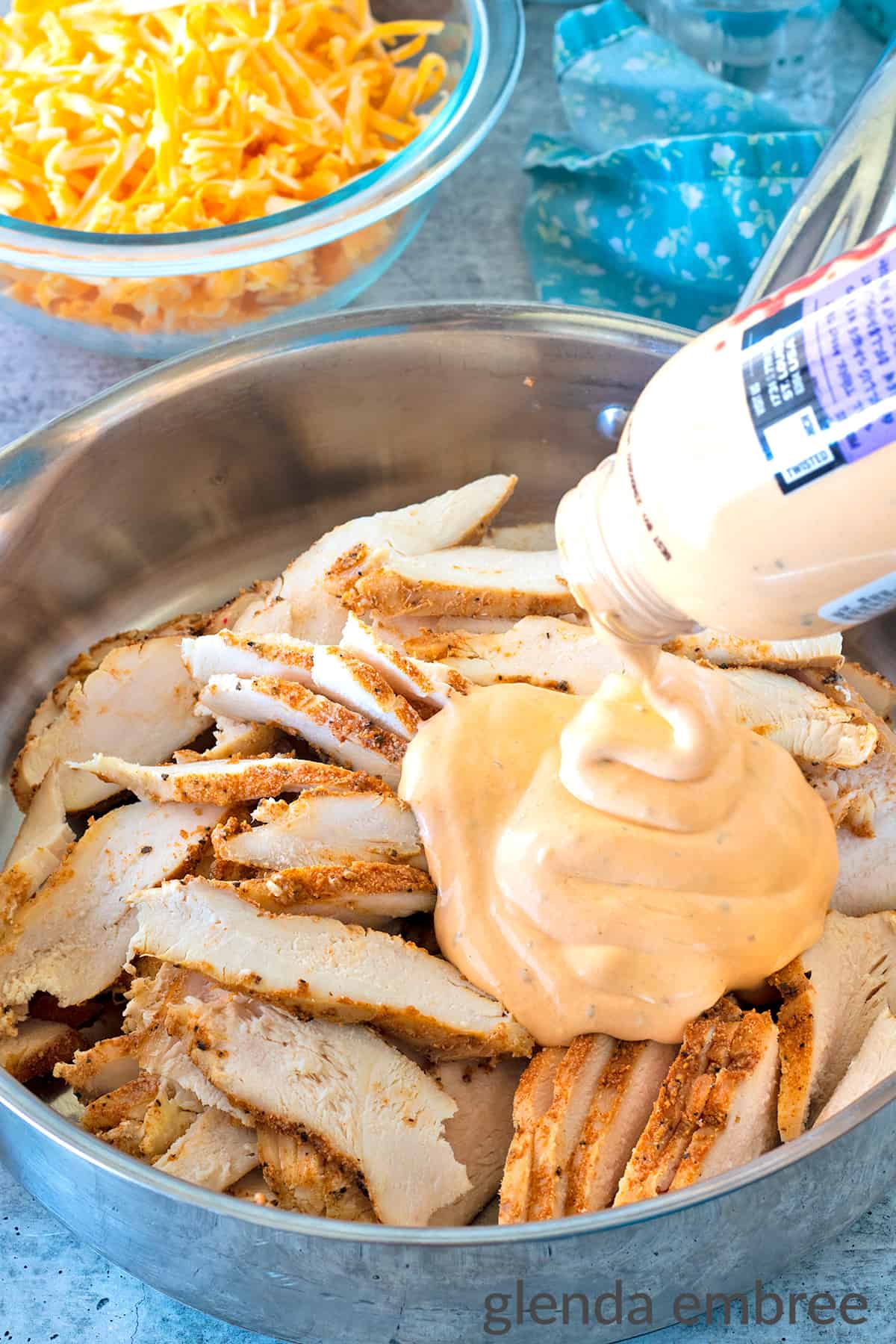 Thinly sliced pre-cooked chicken breast in a stainless steel skillet with buffalo ranch sauce being added.