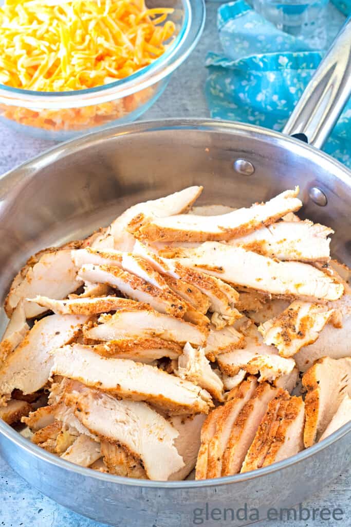 Thinly sliced chicken breast in a stainless steel skillet.  Skillet is sitting on a concrete countertop next to a bowl of grated cheese and a blue fabric napkin.