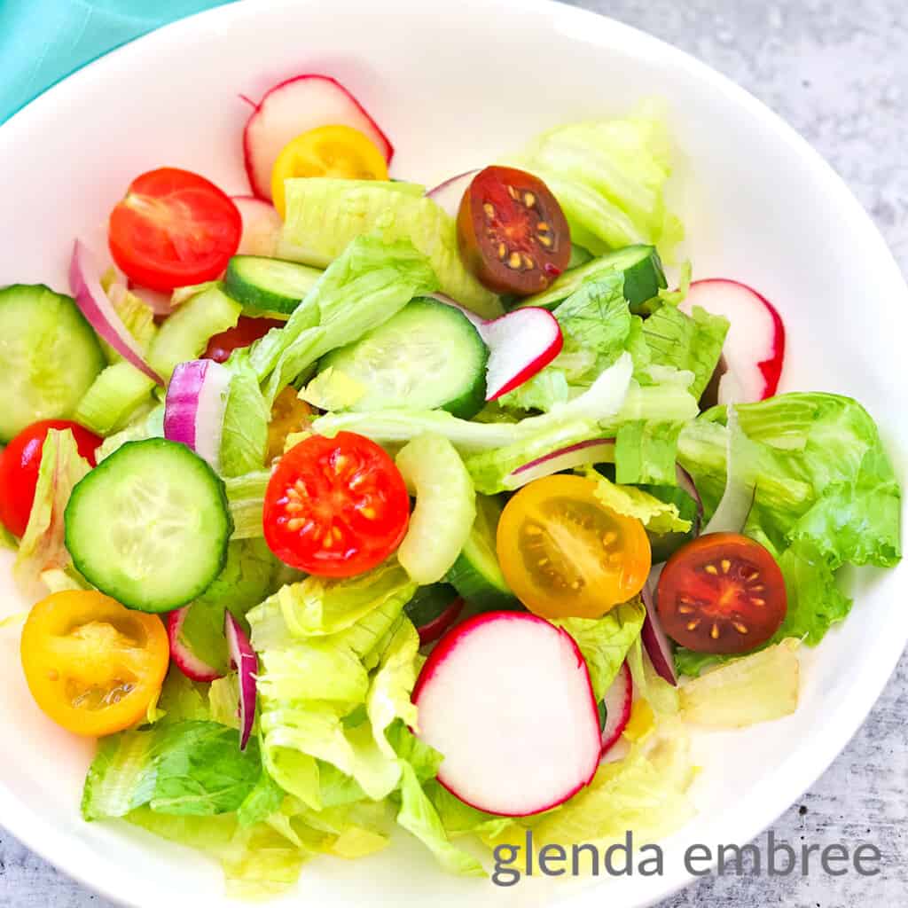 House Salad in a white bowl: salad contains romaine and iceburg lettuce with halved yellow and red cherry tomatoes, radish slices, celery slices, cucumber slices and slivered red onion