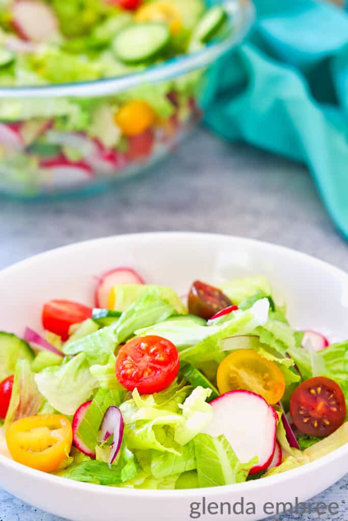 A house salad in a bowl ona concrete counter top with a turquoise fabric napkin. The larger salad bowl of salad is in the background