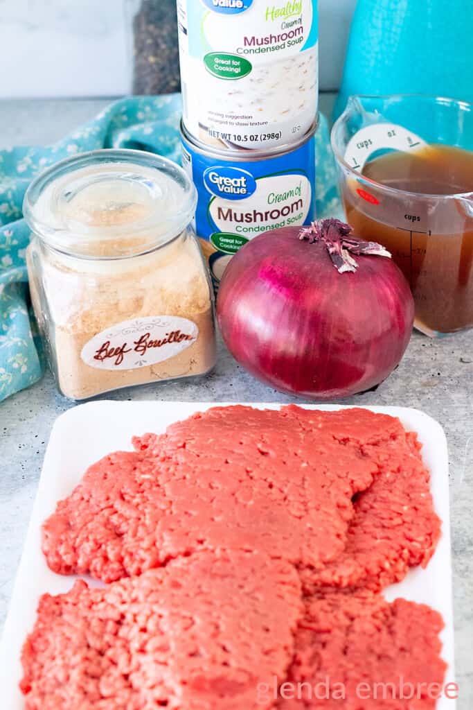 Ingredients for Crock Pot Cube Steak: cube steaks, red onion, bouillon powder, beef broth and cream of mushroom soup.  Ingredients are on a concrete counter next to a blue print fabric napkin