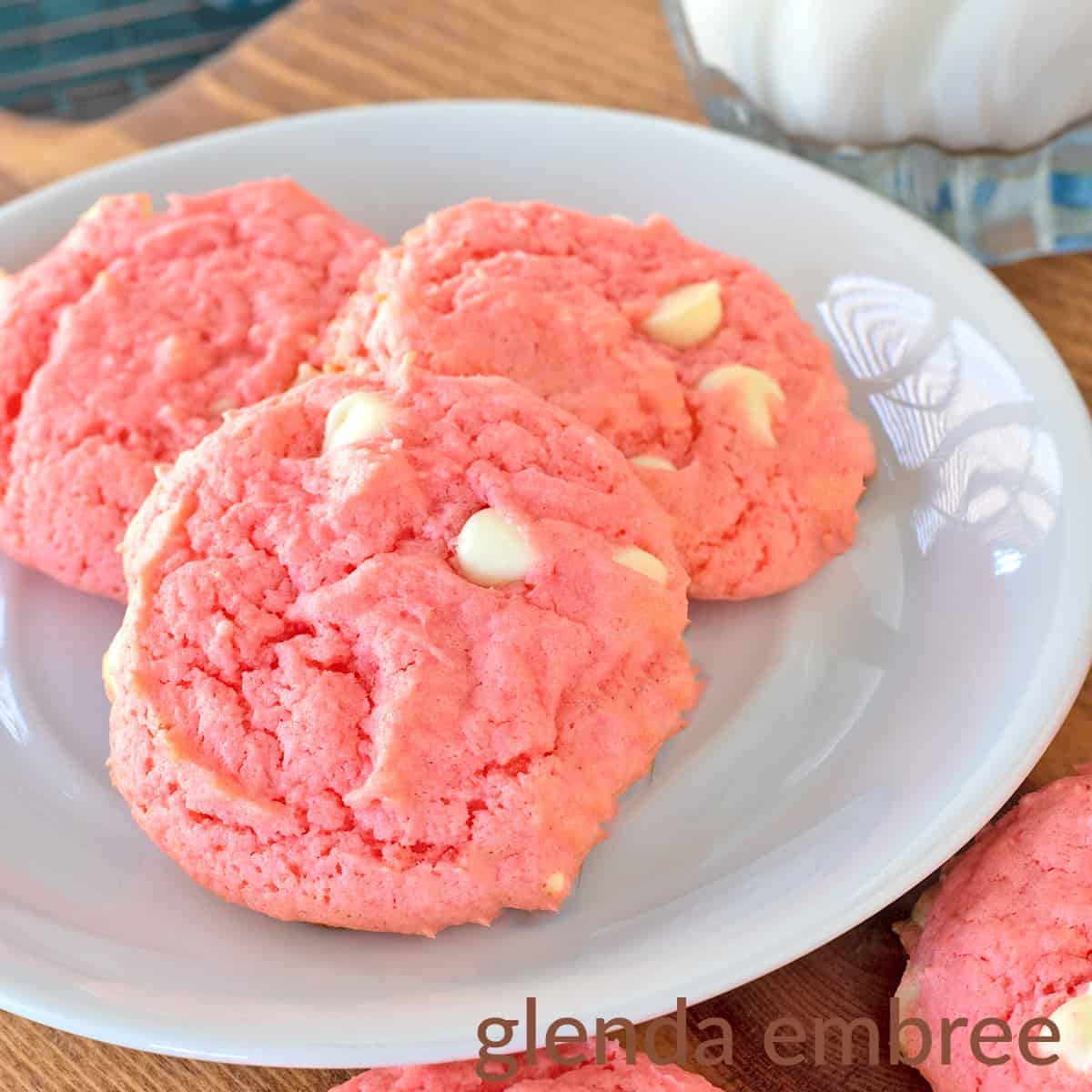 Strawberry Cake Mix Cookies are pink and dotted with white chocolate chips. Cookies and a glass of milk are on a white plate that is resting on a wooden trivet.