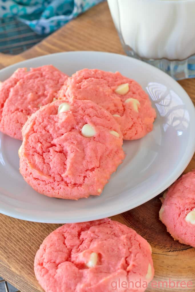 Strawberry Cake Mix Cookies are pink and dotted with white chocolate chips. Cookies and a glass of milk are on a white plate that is resting on a wooden trivet.