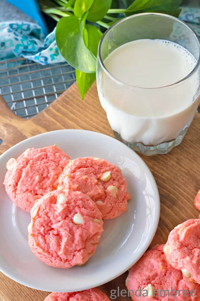 Strawberry Cake Mix Cookies are pink and dotted with white chocolate chips.  Cookies and a glass of milk are on a white plate that is resting on a wooden trivet.