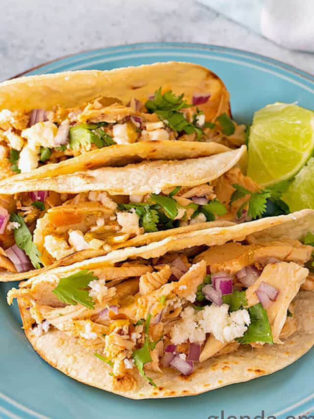 Three Chicken Street Tacos on a blue stoneware plate. Southwest Chicken is placed into charred corn tortillas and topped with minced red onion, chopped cilantro and queso fresco cheese. Fresh lime wedges are tucked on the side for drizzling.