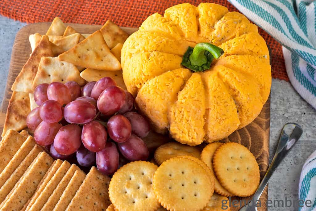 Bacon Pineapple cheeseball on a wooden cutting board with an assortment of crackers and fresh red grapes. Cheeseball is molded into the shape of a pumpkin.