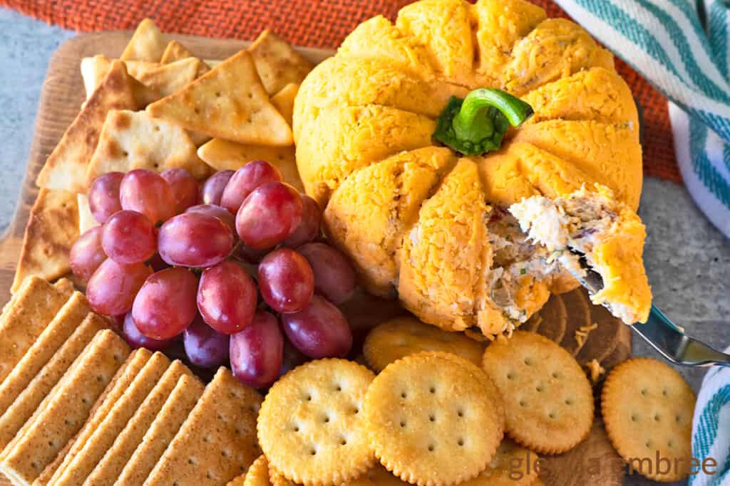 Bacon Pineapple cheeseball on a wooden cutting board with an assortment of crackers and fresh red grapes. Cheeseball is molded into the shape of a pumpkin.  A dab of cheese ball is shown on the end of a cheese spreader.