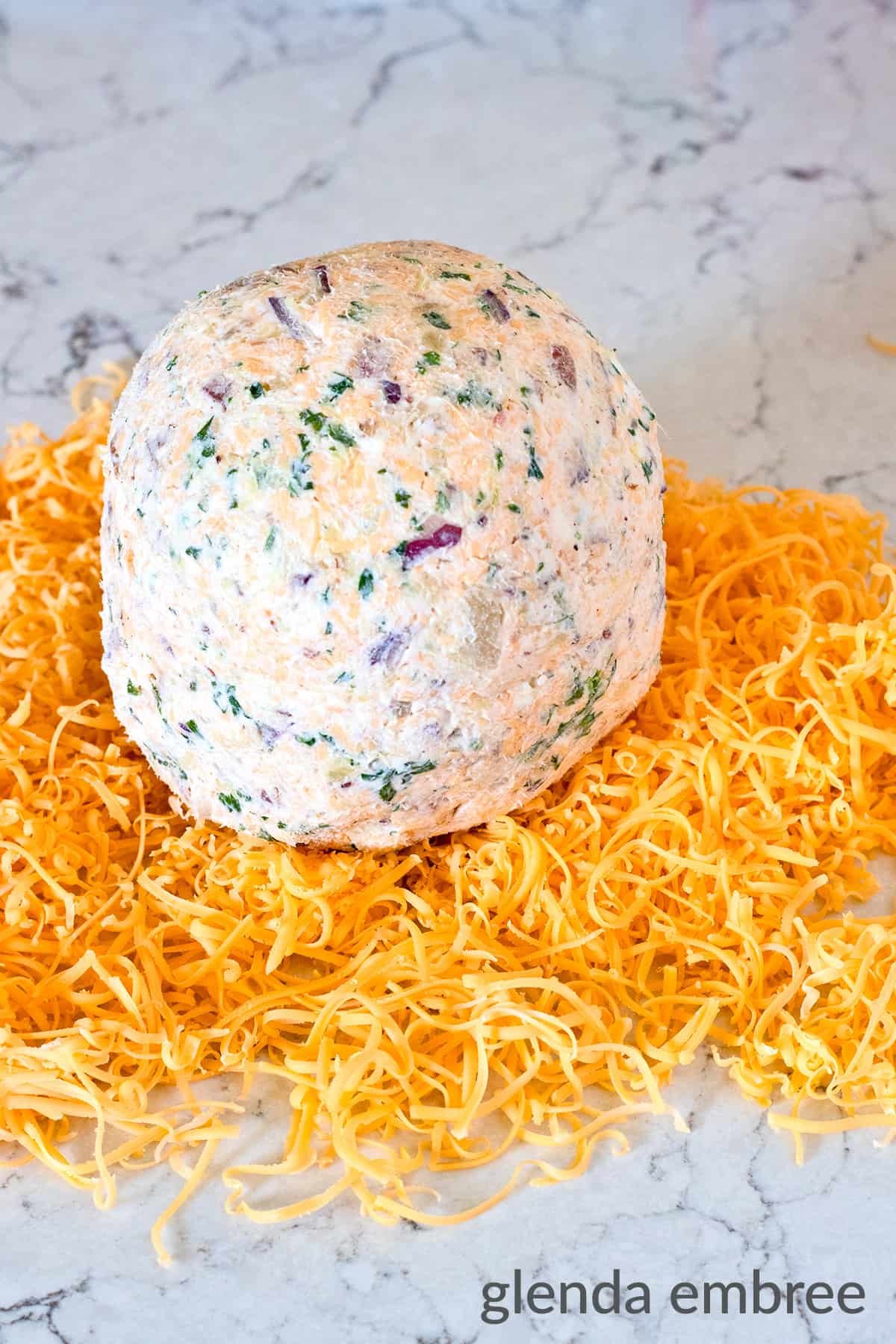 Bacon and Pineapple Cheese Ball in the center of a pile of grated Cheddar cheese on a marble counter