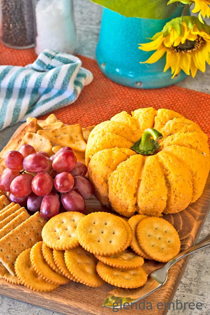 Bacon Pineapple cheeseball on a wooden cutting board with an assortment of crackers and fresh red grapes. Cheeseball is molded into the shape of a pumpkin.