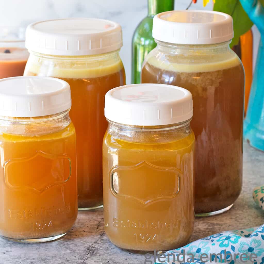 Homemade Chicken Broth on a concrete counter. The broth is in 2 quart jars and 2 pint jars sitting in front of an arrangement of sunflowers