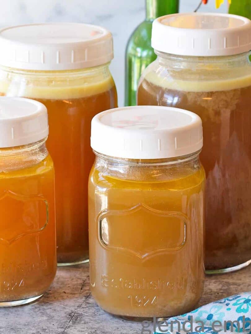 Homemade Chicken Broth on a concrete counter. The broth is in 2 quart jars and 2 pint jars sitting in front of an arrangement of sunflowers