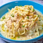 Creamy Chicken and Noodles over Mashed Potatoes in a blue stoneware bowl on a concrete counter. A turquoise pitcher with sunflowers and a turquoise and white striped tea towel are in the background.