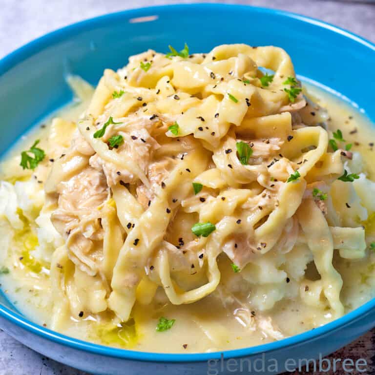 Creamy Chicken and Noodles over Mashed Potatoes in a blue stoneware bowl on a concrete counter. A turquoise pitcher with sunflowers and a turquoise and white striped tea towel are in the background.