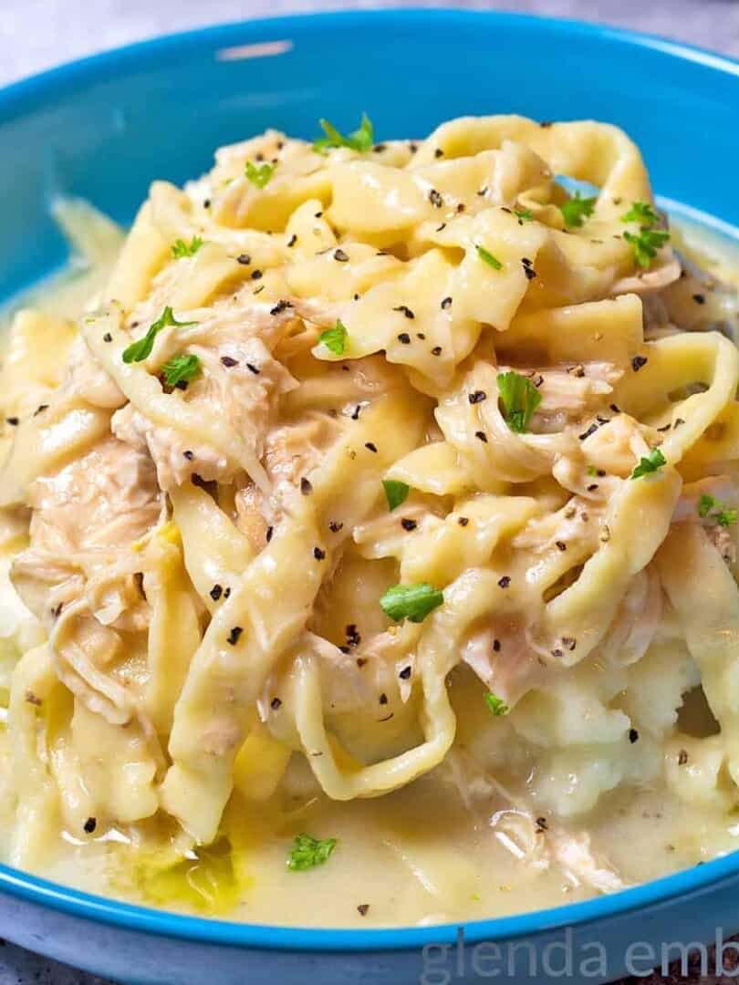 Creamy Chicken and Noodles over Mashed Potatoes in a blue stoneware bowl on a concrete counter. A turquoise pitcher with sunflowers and a turquoise and white striped tea towel are in the background.