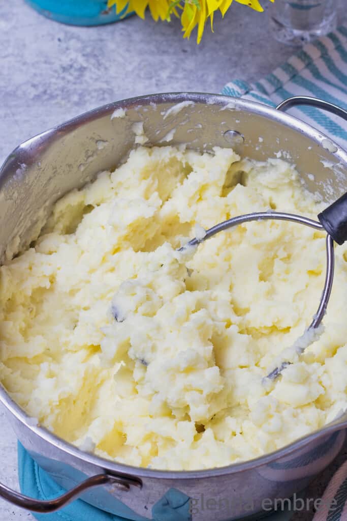 Mashed potatoes in a stainless steel pot with a potato masher.
