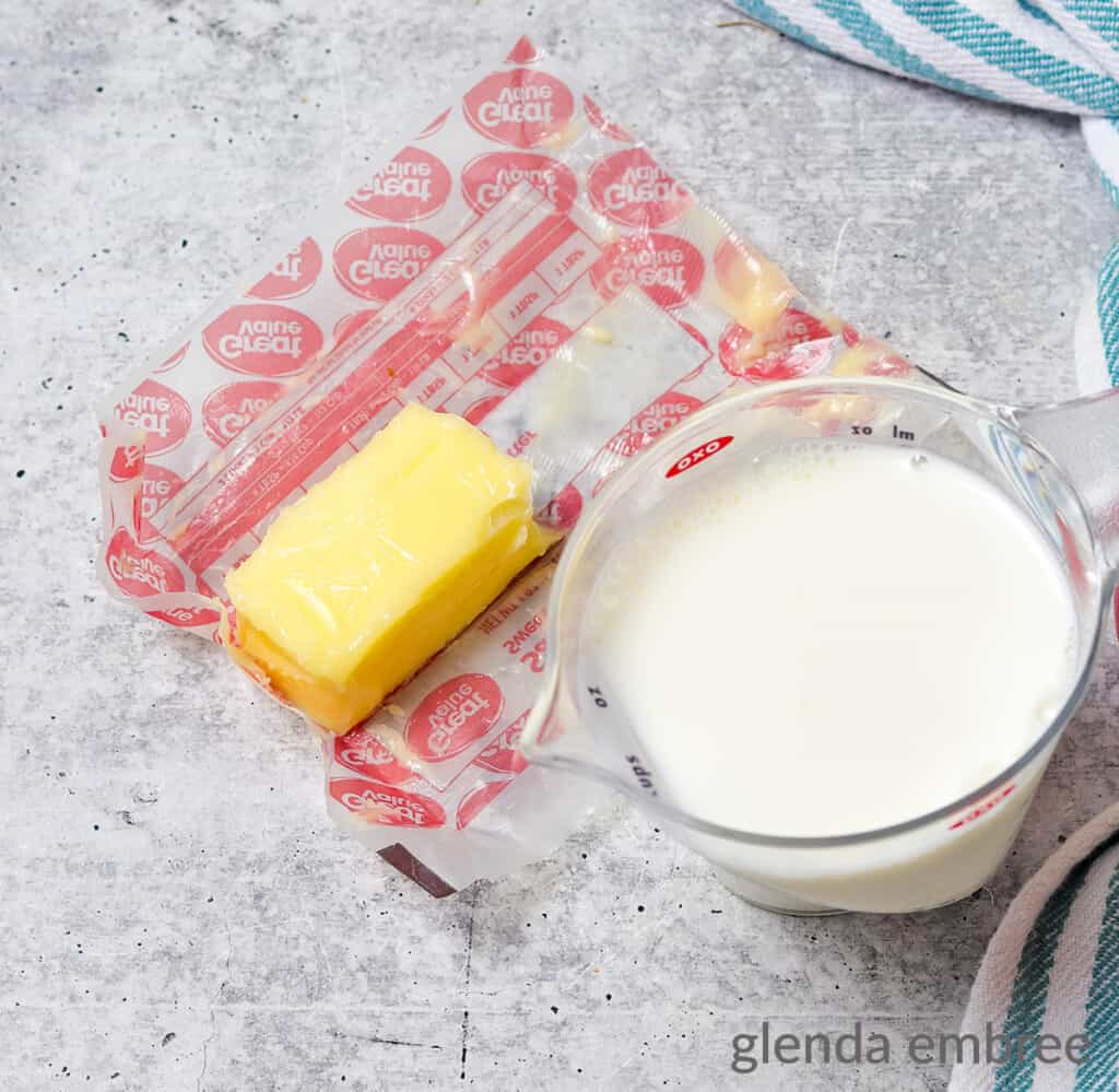 One cup of whole milk measured into a clear plastic measuring cup and sitting next to half a stick of softened butter