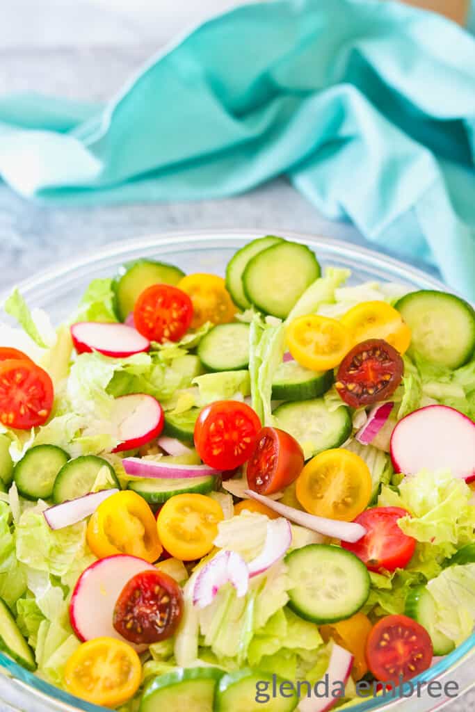 Clear glass serving bowl foull of Classic House Salad with lettuce, tomatoes, celery, radishes, cucumber and slivered red onions