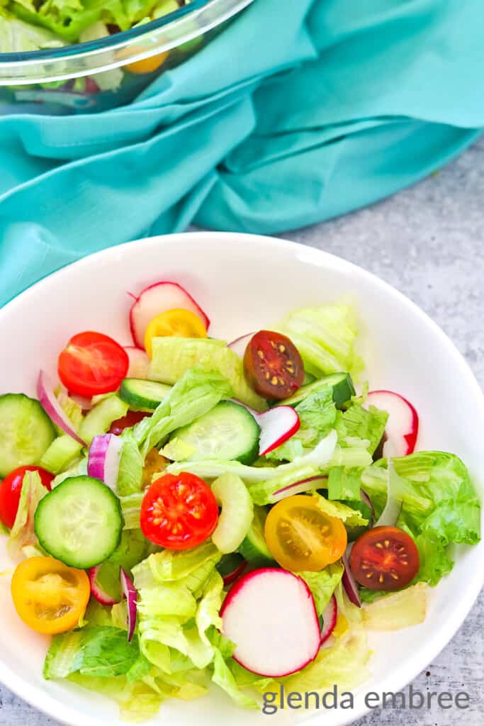 A house salad in a bowl ona concrete counter top with a turquoise fabric napkin. The larger salad bowl of salad is in the background