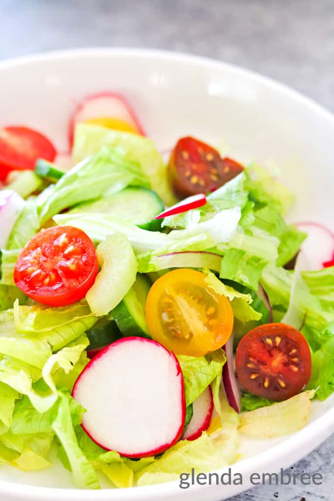 House Salad in a white ceramic bowl on a concrete countertop. There is a fork lying next to the bowl witha turquoise fabric napkin.