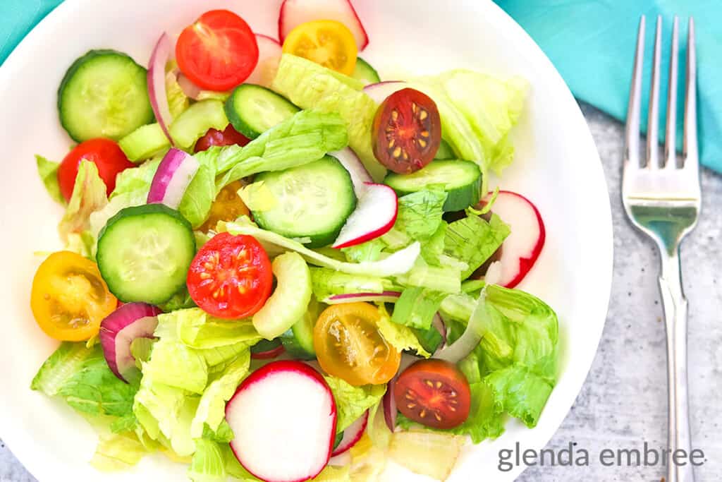 House Salad in a white ceramic bowl on a concrete countertop. There is a fork lying next to the bowl witha turquoise fabric napkin.