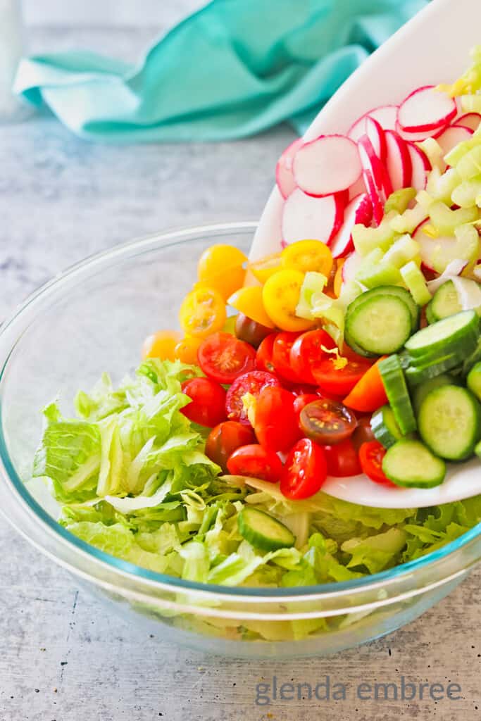 Adding chopped vegetables to salad greens in a clear mixing bowl on a concrete countertop