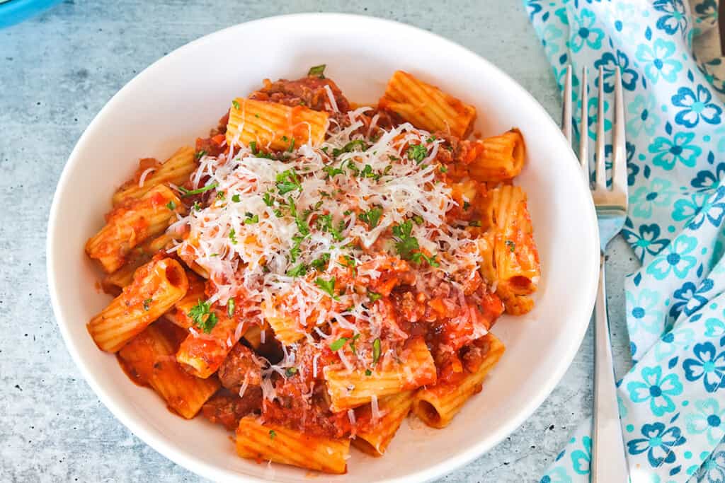 Rigatoni Bolognese served in a white stoneware bowl with low sides. A blue print fabric napkin and a fork are to the right of the bowl.