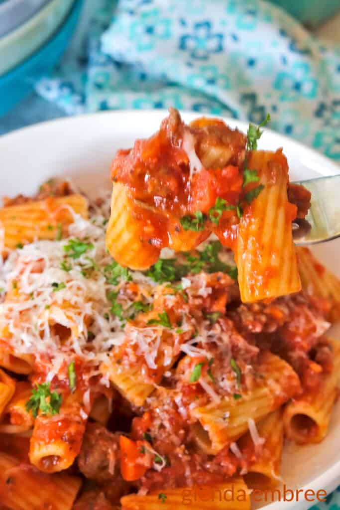 Rigatoni Bolognese served in a white stoneware bowl with low sides. A blue print fabric napkin is in the background and a bite is being lifted out of the bowl on a fork.
