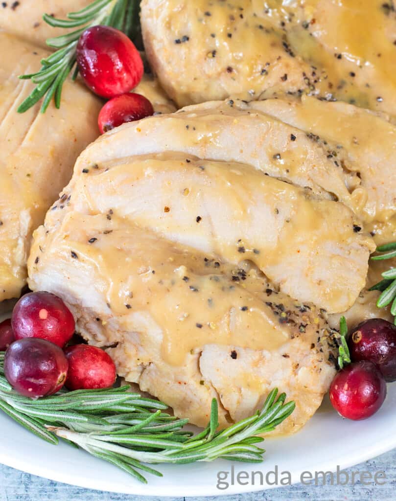 crockpot turkey tenderloins on a white platter surrounded by fresh rosemary and cranberries. platter is on a concrete countertop with a blue print fabric napkin and a white gravy boat filled with turkey gravy