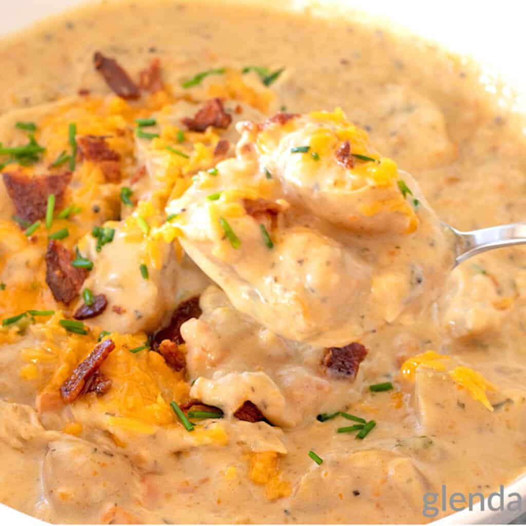 Crockpot Potato Soup garnished with grated cheddar, bacon and chives. Soup is served in a white ceramic bowl on a concrete countertop with a blue and white print fabric napkin and a buttered biscuit sitting nearby.