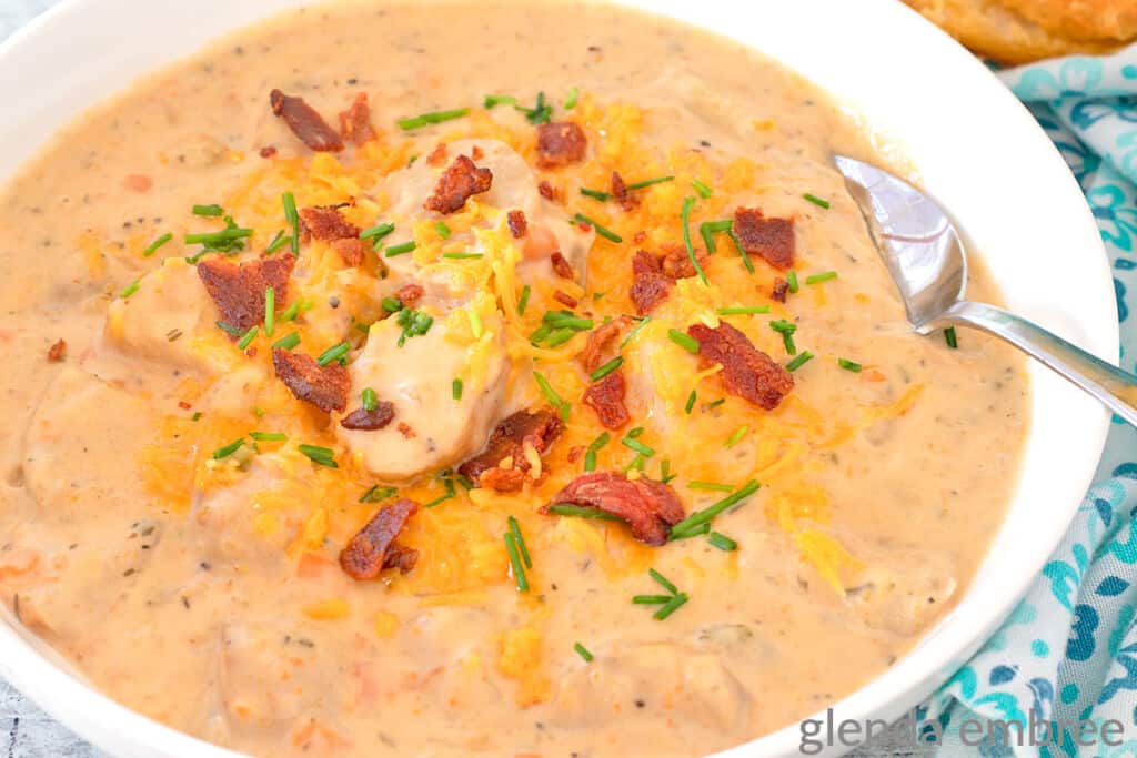 Crockpot Potato Soup garnished with grated cheddar, bacon and chives. Soup is served in a white ceramic bowl on a concrete countertop with a blue and white print fabric napkin and a buttered biscuit sitting nearby.