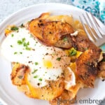 Fried Potatoes and Onions on a White plate. The potatoes are served with a fried egg on top. Plate is on a concrete countertop with a fork and blue print napkin.