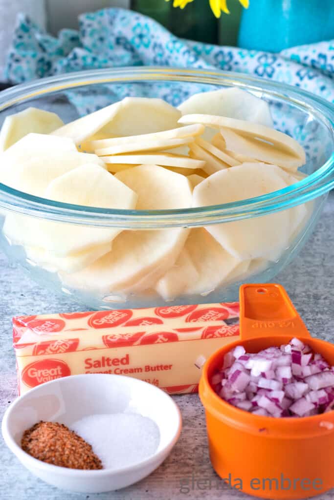 Ingredients for Fried Potatoes and Onions on a Concrete Counter top:  Clear bowl of peeled and sliced Russet Potatoes, stick of softened butter, 1/2 cup of minced red onion and small white bowl with All Purpose Seasoning and Salt in it.