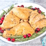 crockpot turkey tenderloins on a white platter surrounded by fresh rosemary and cranberries. platter is on a concrete countertop with a blue print fabric napkin and a white gravy boat filled with turkey gravy
