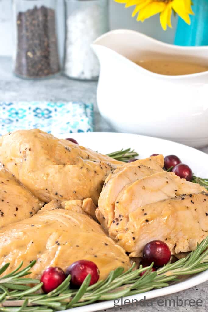 crockpot turkey tenderloins on a white platter surrounded by fresh rosemary and cranberries. platter is on a concrete countertop with a blue print fabric napkin and a white gravy boat filled with turkey gravy