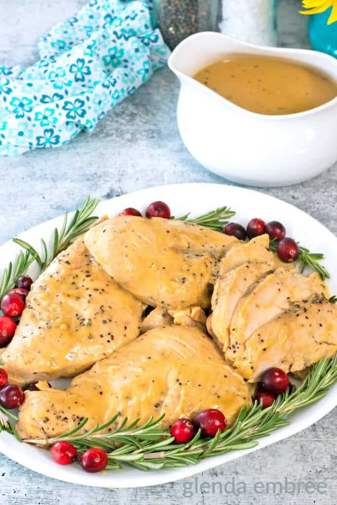 Crockpot turkey tenderloins on a white platter with fresh rosemary and cranberries.  Platter is on a concrete countertop with a white gravy boat of turkey gravy and a blue and white print fabric napkin
