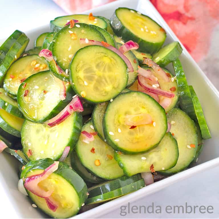 Asian Cucumber Salad served in a square, white bowl sitting on a marble countertop. A watercolor print napkin is to the right of the bowl and a turquoise planter is in the background.