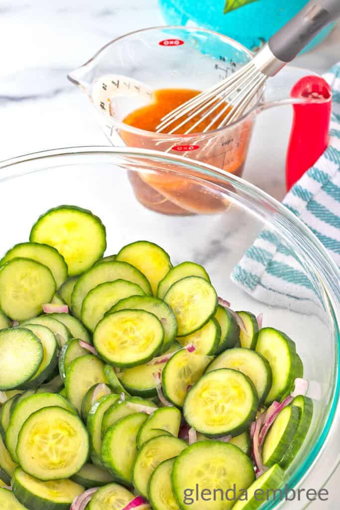 Prepped cucumber and onion slices in a clear glass bowl. Dressing for Asian Cucumber Salad whisked in a measuring cup behind the cucumber bowl.