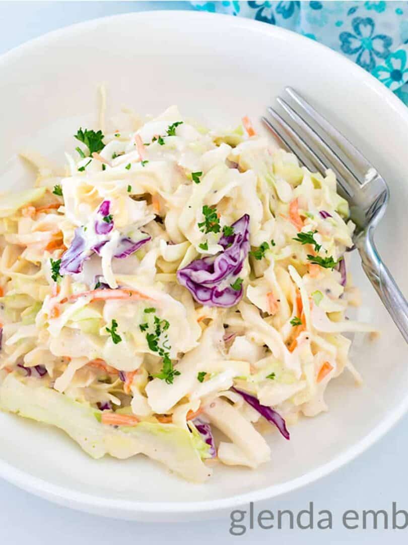 Best Easy Coleslaw Recipe served in a white bowl on a marble countertop. A blue and white print fabric napkin is in the background.